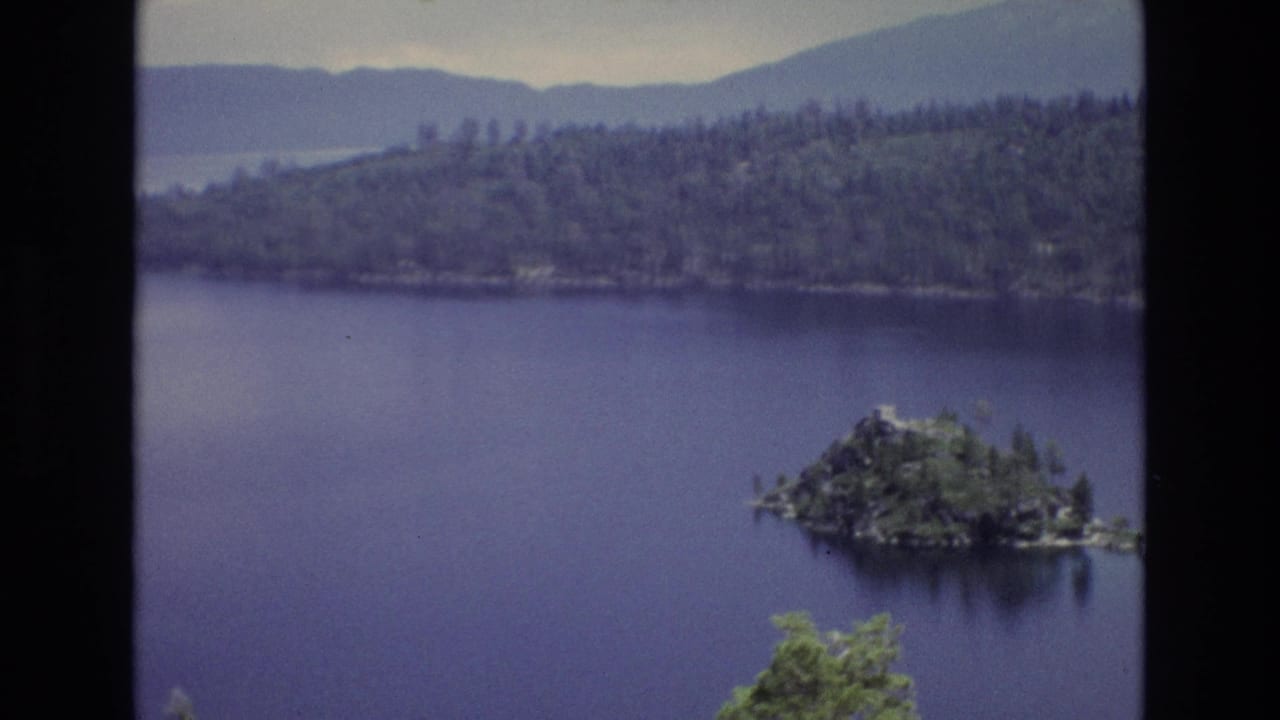 Lake view with small wooded island near the right side, forested shore, mountains, and dark vertical frame border.