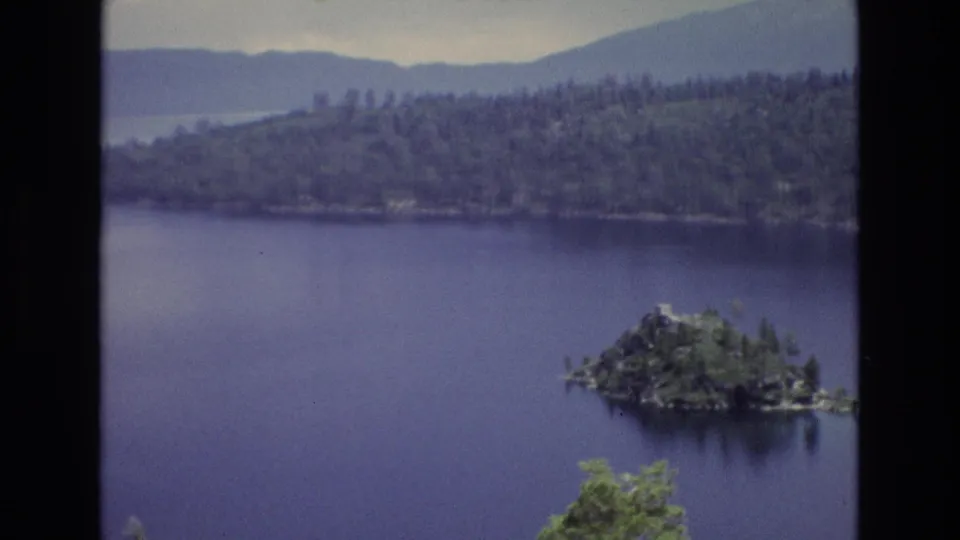1973 Lake Tahoe home-movie frame showing a small wooded island on blue water, forested shoreline, distant mountain ridges, and a prominent dark 8mm frame edge.
