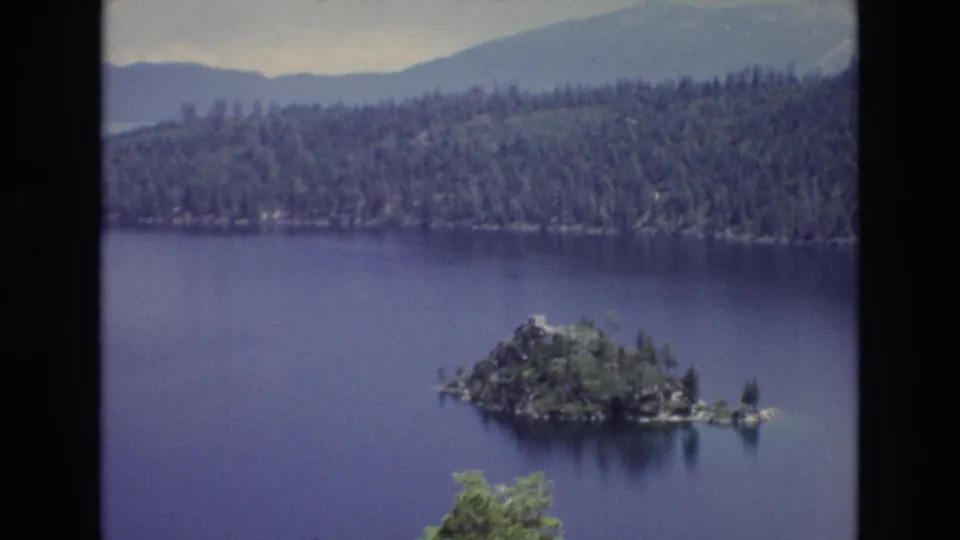 1973 8mm Lake Tahoe frame with the wooded island more centered against blue water, a dense pine shoreline, distant mountains, and faded home-movie color.