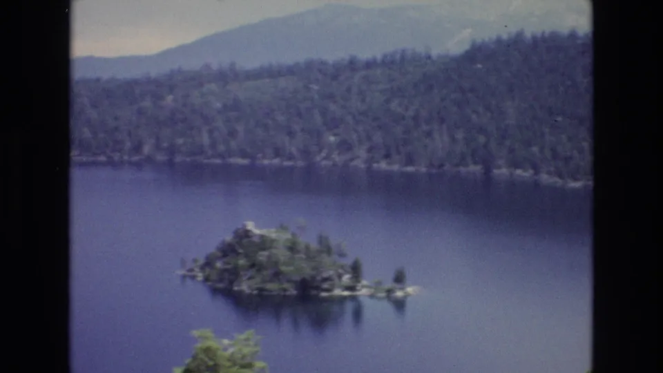 Late-frame 1973 Lake Tahoe view showing the small wooded island on deep blue water, forested shoreline and hills beyond, with soft grain and a dark 8mm border.
