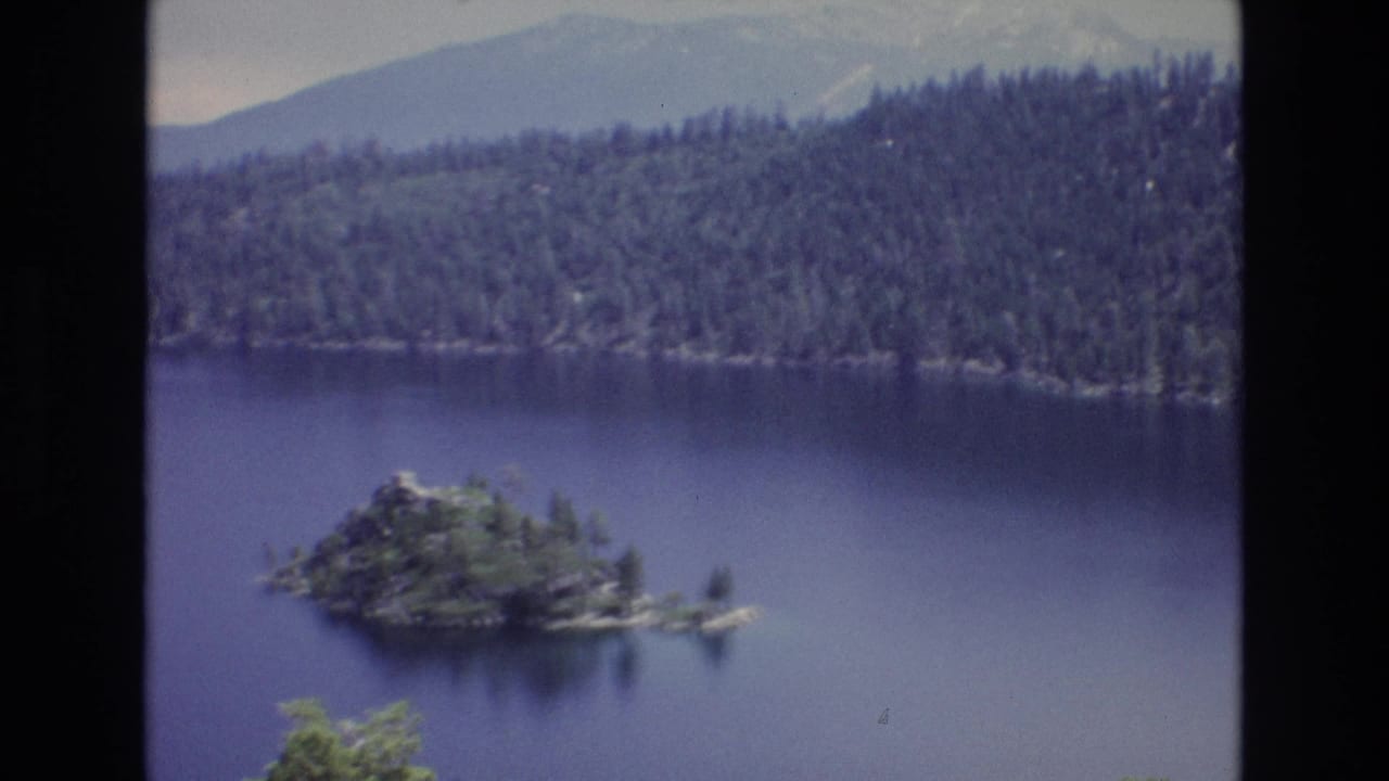1973 8mm home-movie frame of Lake Tahoe, Nevada, showing a small wooded island in deep blue water with evergreen forest and hazy mountain ridges in the background.