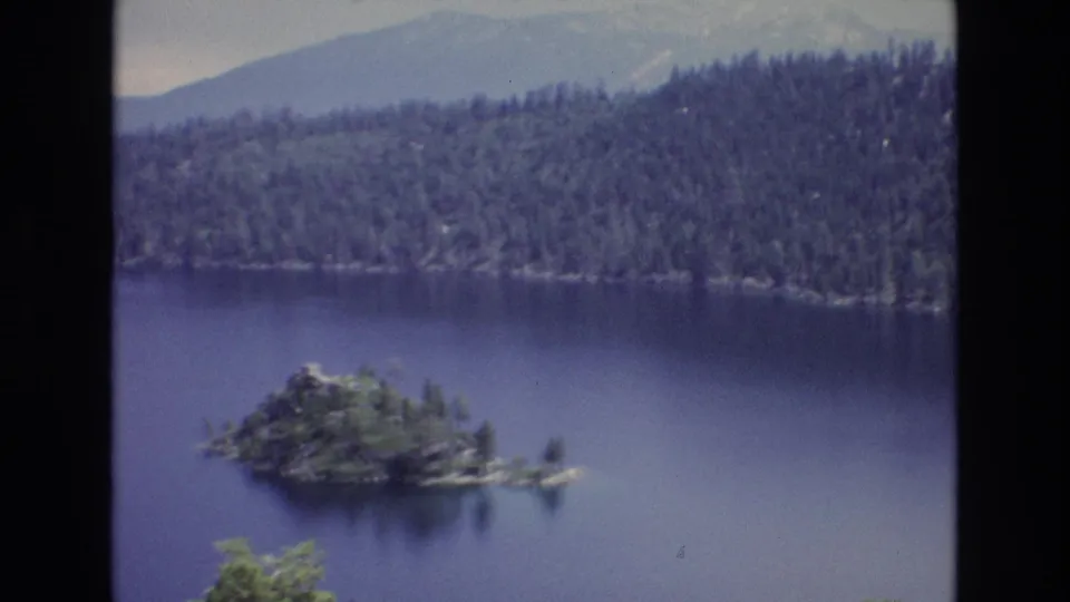 1973 8mm home-movie frame of Lake Tahoe, Nevada, showing a small wooded island in deep blue water with evergreen forest and hazy mountain ridges in the background.