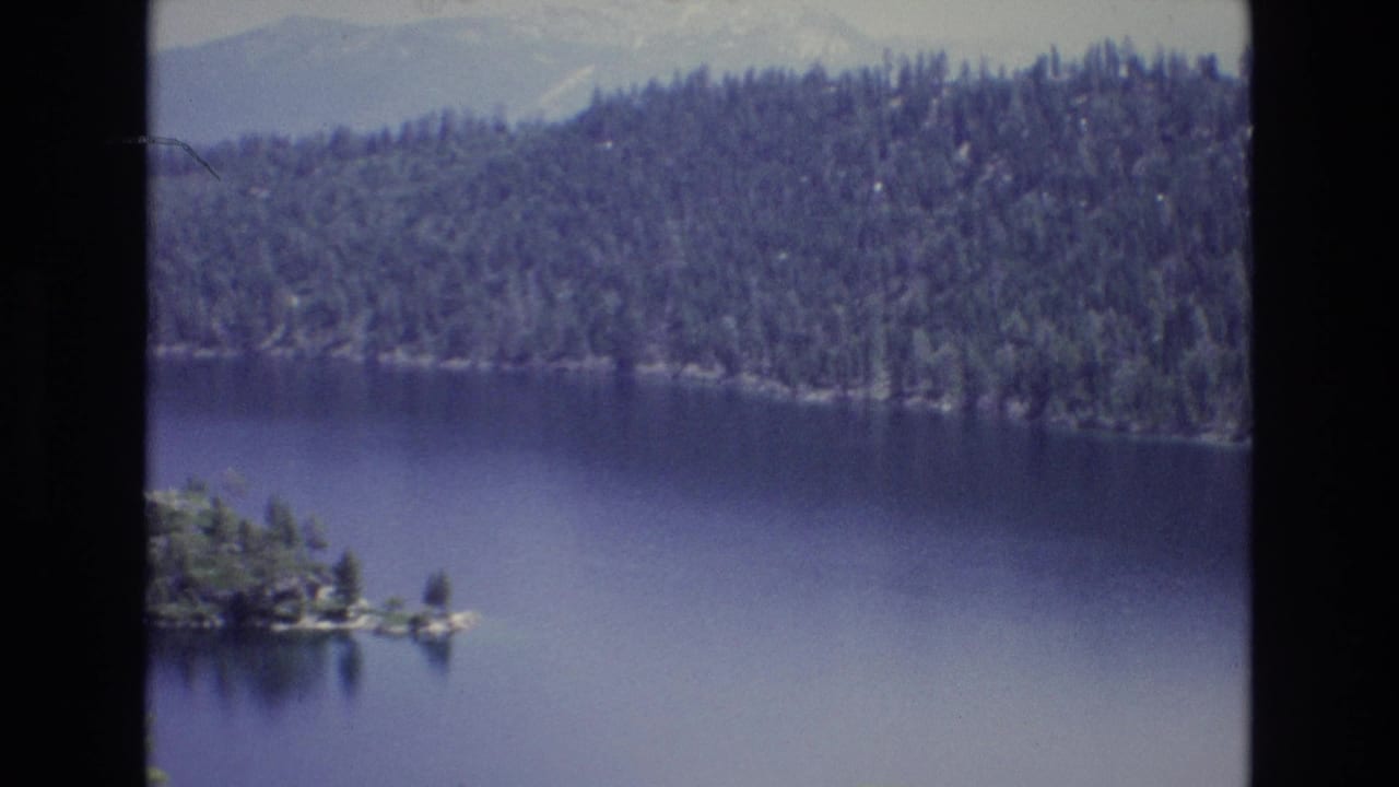 1973 Lake Tahoe home-movie frame with the camera panned right, keeping the forested shoreline and distant mountain ridge in view while the island slips toward the left edge.