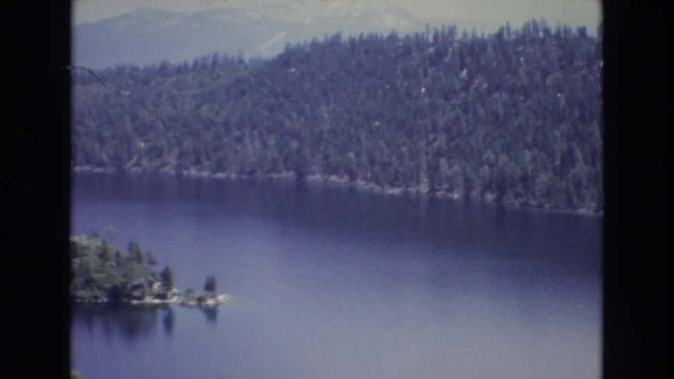 1973 Lake Tahoe home-movie frame with the camera panned right, keeping the forested shoreline and distant mountain ridge in view while the island slips toward the left edge.