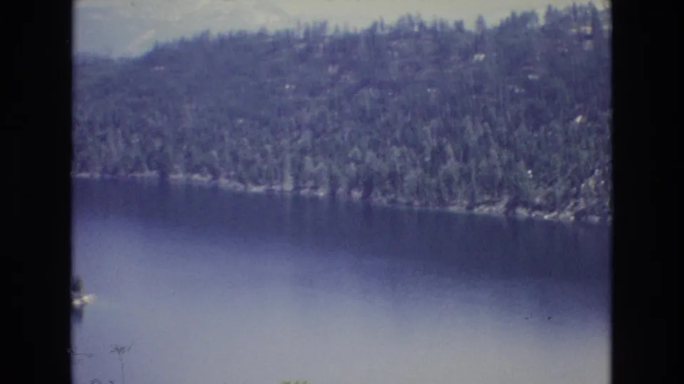 1973 8mm frame from Lake Tahoe, Nevada, showing open blue water in the foreground and a wooded shoreline with hazy hills or mountains behind it.