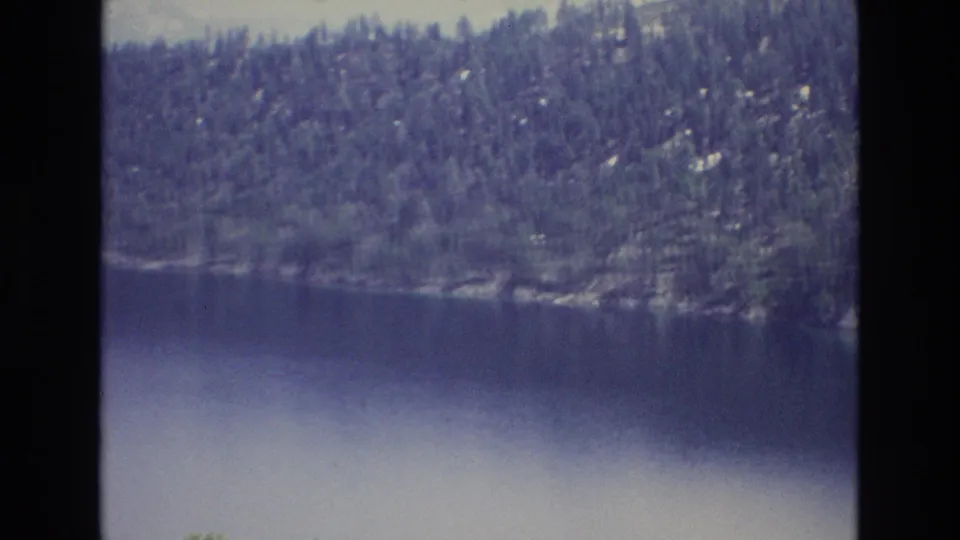 1973 Lake Tahoe 8mm home-movie frame showing calm blue water, a continuous evergreen shoreline, and a hazy mountain ridge under a pale sky.