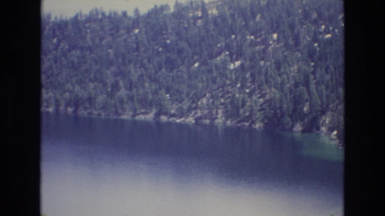 1973 Lake Tahoe home-movie frame showing deep blue water below a densely forested rocky mountain shore.