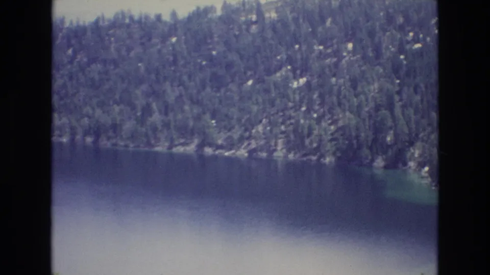1973 Lake Tahoe home-movie frame showing deep blue water below a densely forested rocky mountain shore.