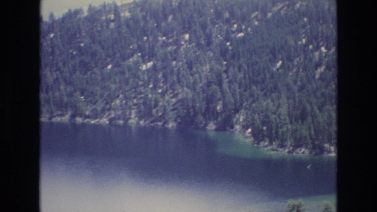 1973 Lake Tahoe home-movie frame with calm blue water, rocky shoreline, and dense evergreen forest on the mountainside.