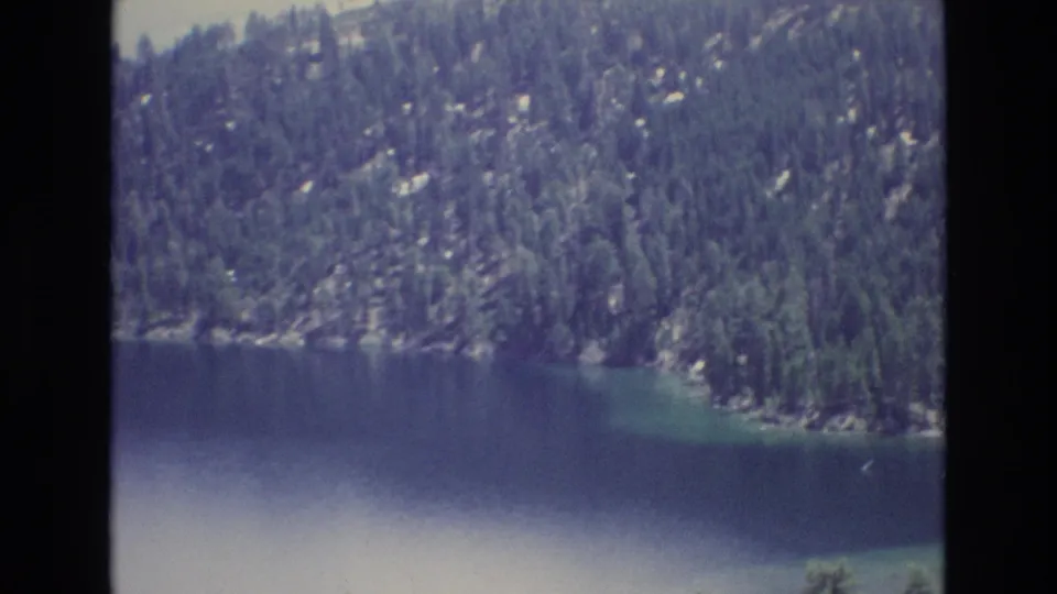 1973 Lake Tahoe home-movie frame with calm blue water, rocky shoreline, and dense evergreen forest on the mountainside.