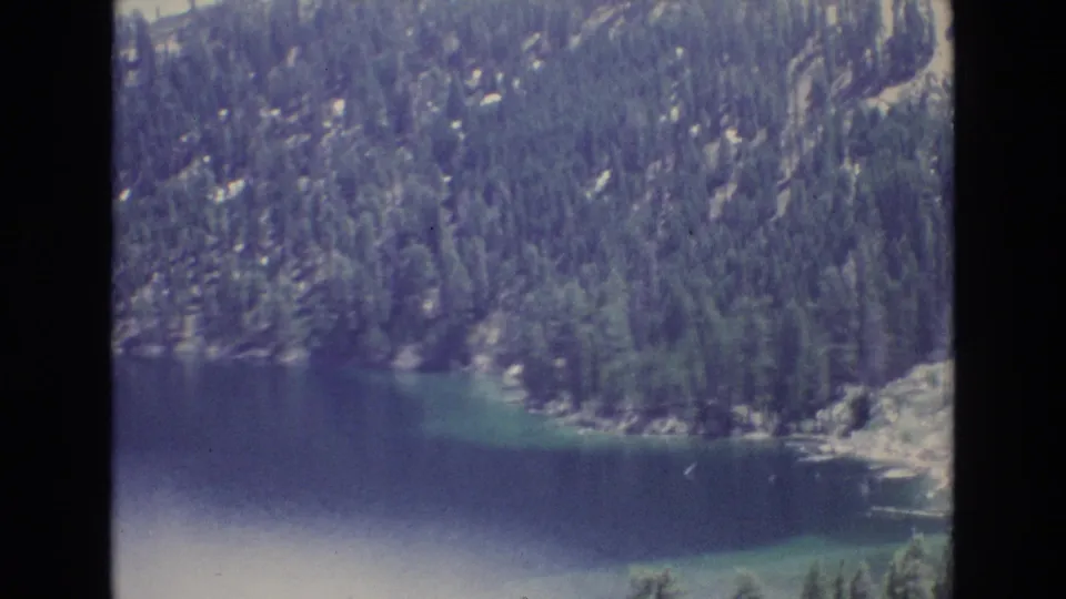 1973 Lake Tahoe frame showing a wooded shoreline curving along dark blue water with rocks and evergreen trees on the slope.