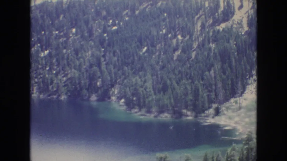 1973 Lake Tahoe home-movie frame with calm lake water, green shallows, rocky shore, and dense evergreen trees on the mountainside.