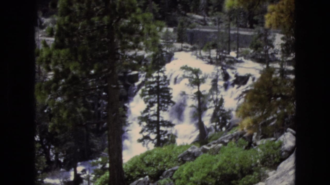 Waterfall partly seen through pine trees in a forested rocky area.