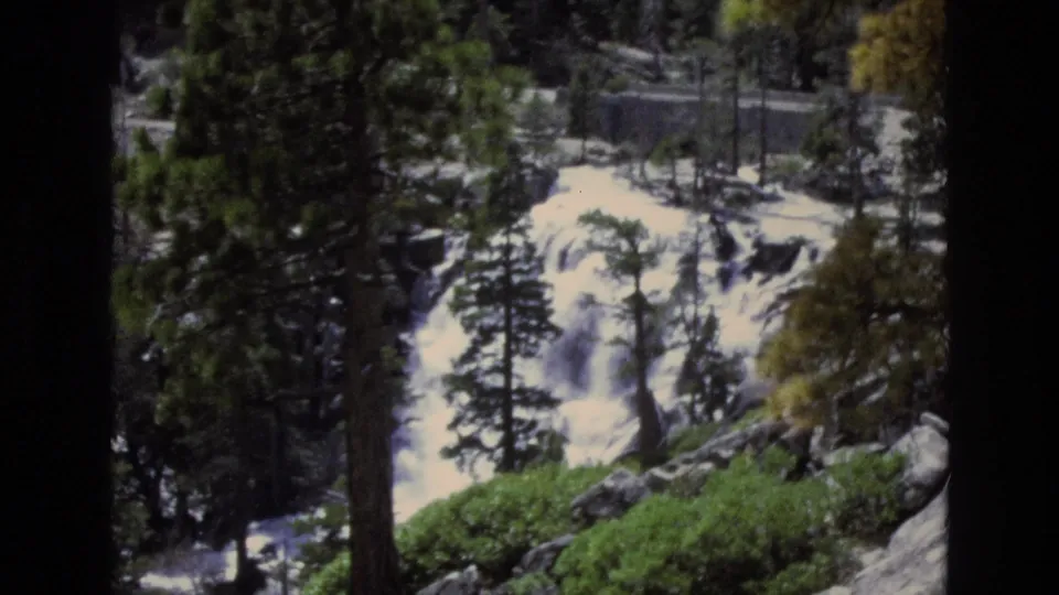 1973 8mm Lake Tahoe, Nevada frame showing white water dropping over rocks behind pine trunks and green undergrowth.