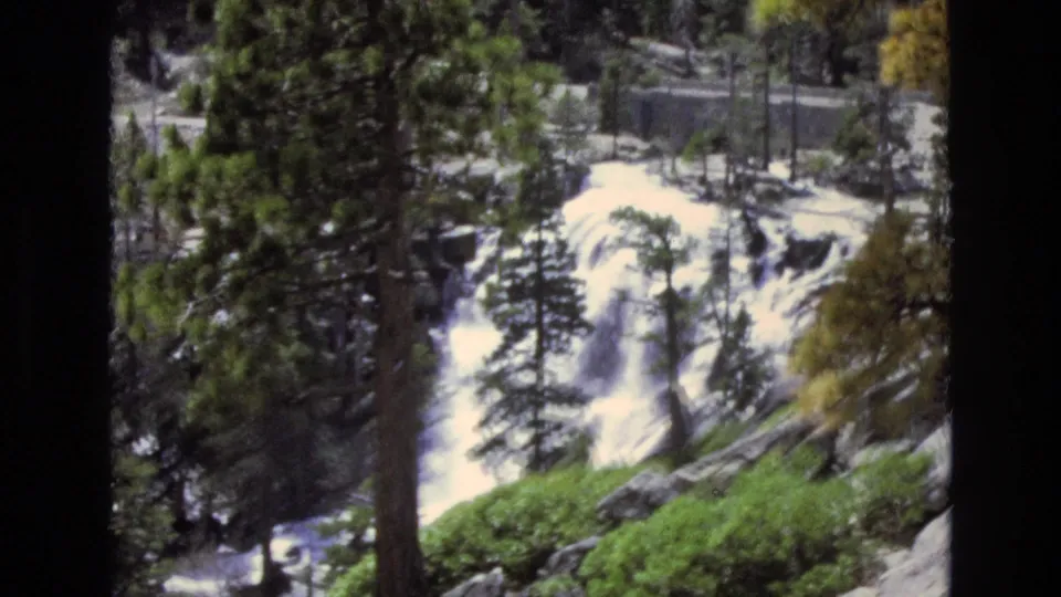 1973 8mm Lake Tahoe, Nevada frame showing a waterfall descending through a pine forest, with a dark trunk interrupting the home-movie view.