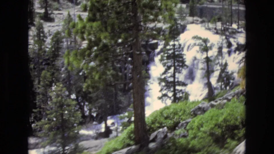 1973 8mm Lake Tahoe, Nevada frame showing rushing white water over rocks, framed by pine trees, shrubs, and a dark foreground trunk.