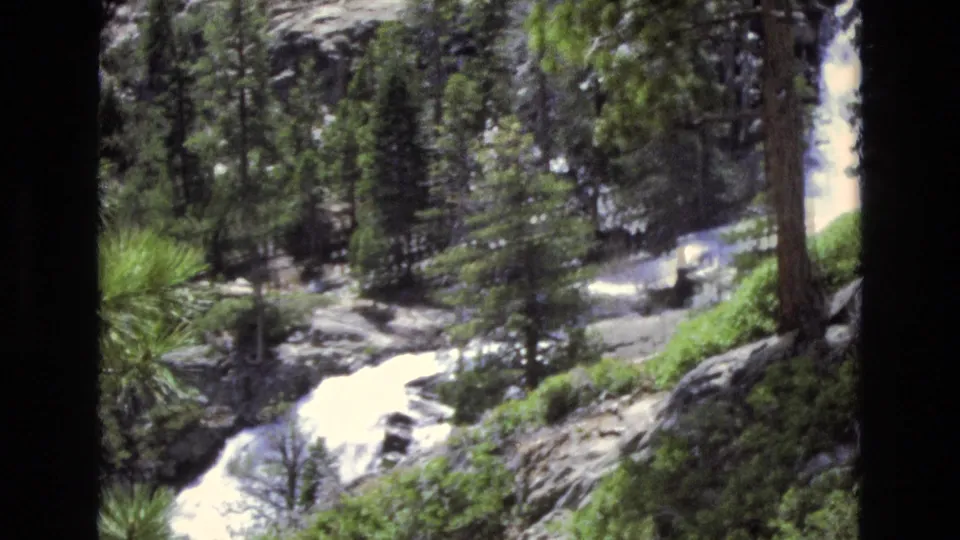 1973 Lake Tahoe, Nevada 8mm frame showing rushing white water, dark pine trunks, rocks, and patches of green brush in a forested ravine.