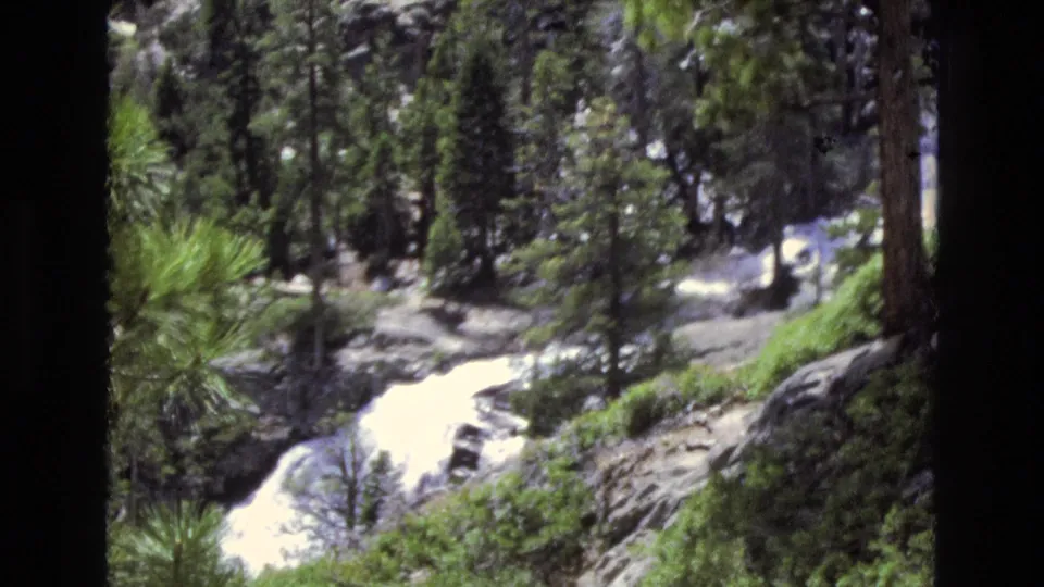 1973 Lake Tahoe, Nevada 8mm frame capturing a rocky stream and waterfall area framed by tall pines, shrubs, and bright daylight on the water.