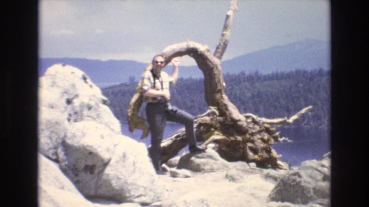 1973 Lake Tahoe, Nevada home-movie frame showing a white adult man in sunglasses and a plaid shirt posing on pale rocks beside a gnarled tree root, with lake water, pine forest, and distant mountains behind him.