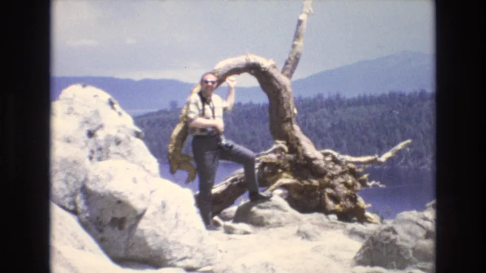 1973 Lake Tahoe, Nevada home-movie frame showing a white adult man in sunglasses and a plaid shirt posing on pale rocks beside a gnarled tree root, with lake water, pine forest, and distant mountains behind him.