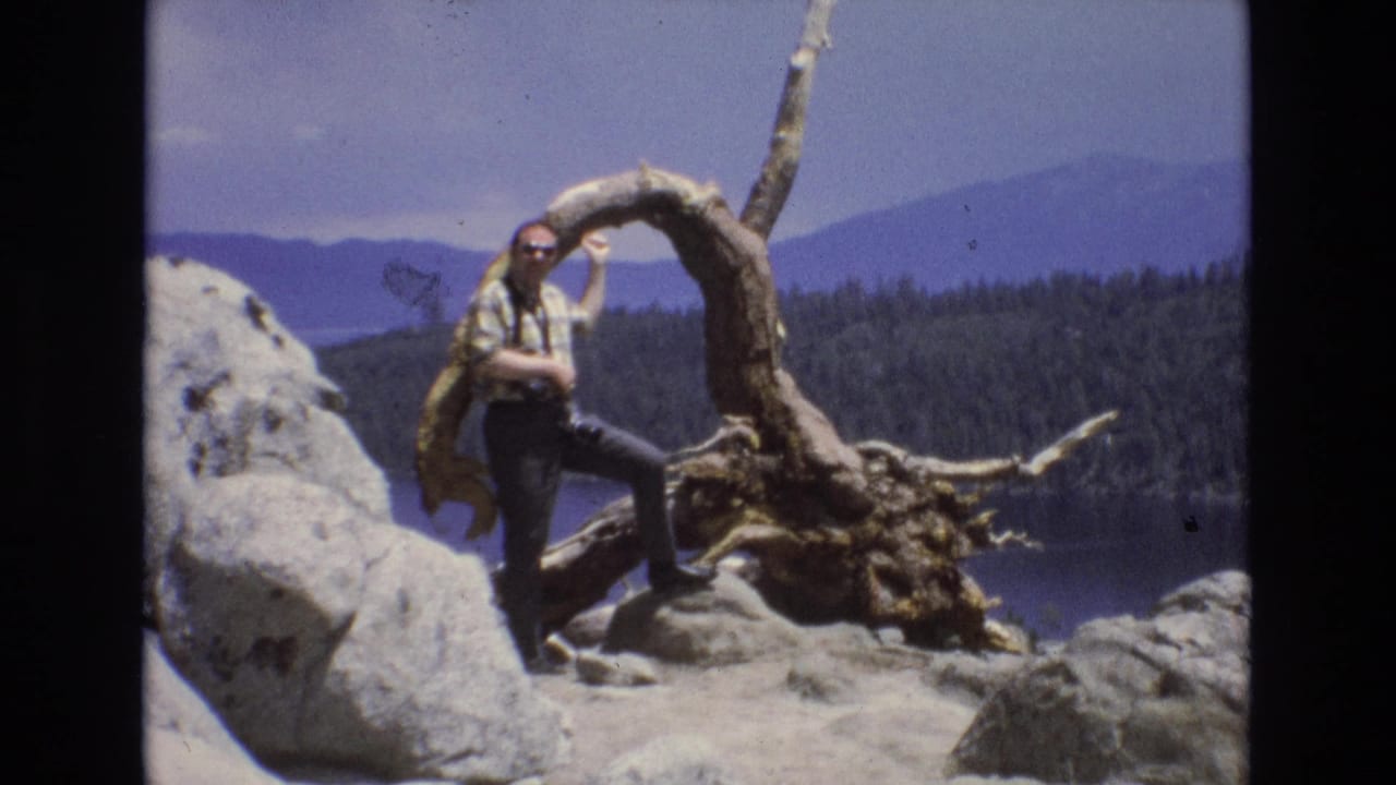 1973 Lake Tahoe, Nevada frame with the man holding a curved exposed tree root while standing on rocky ground, backed by blue lake water, dark forest, and hazy mountain ridges.