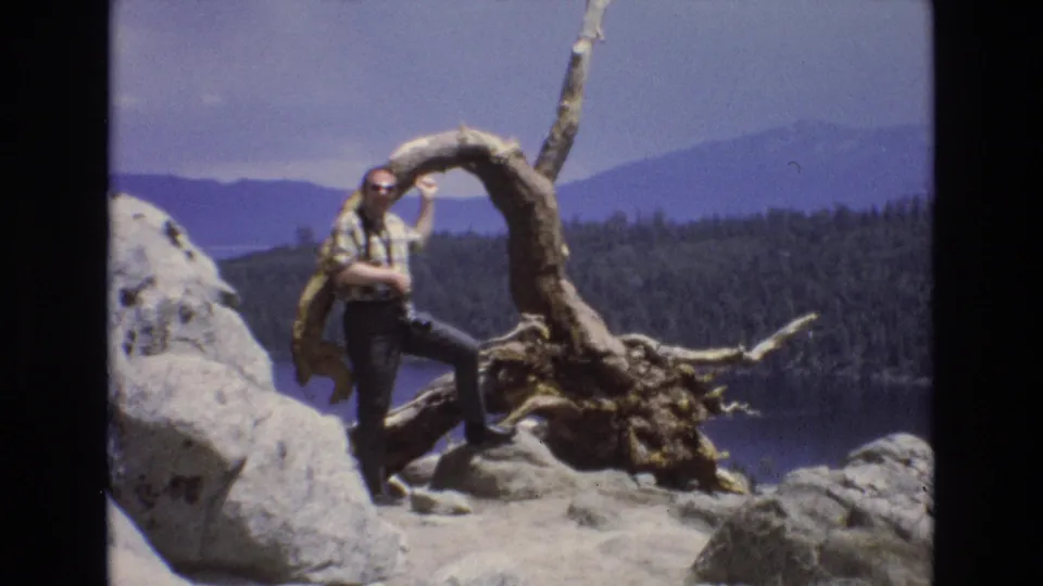 1973 Lake Tahoe, Nevada home-movie still showing the man in a plaid shirt and sunglasses beside a weathered tree root on pale rocks, with forested shoreline and mountains across the lake.