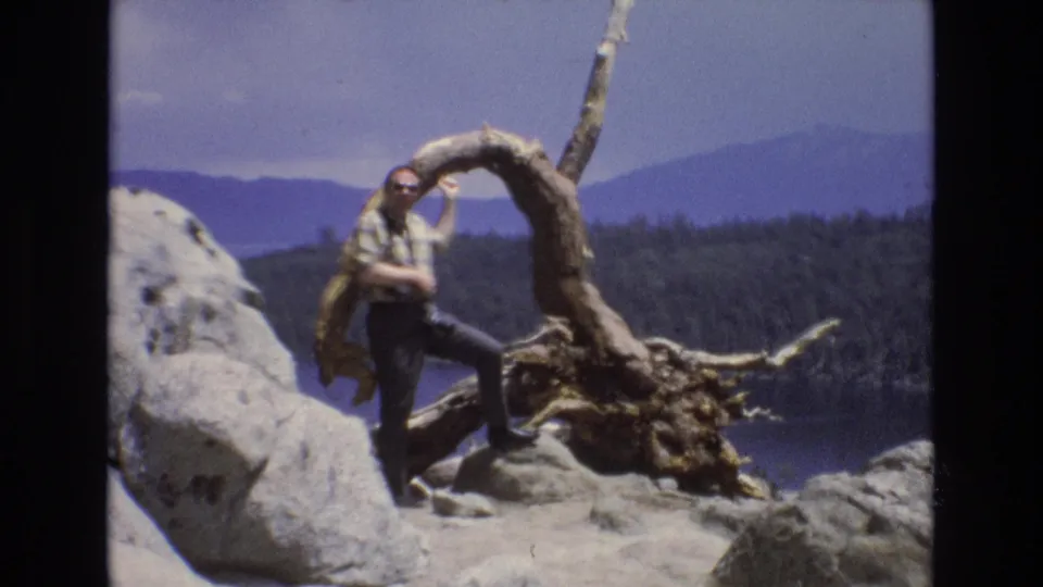 1973 Lake Tahoe, Nevada frame showing the same adult traveler posed next to a gnarled exposed root on a rocky overlook, with the lake, pine-covered land, and blue mountain slopes visible behind him.