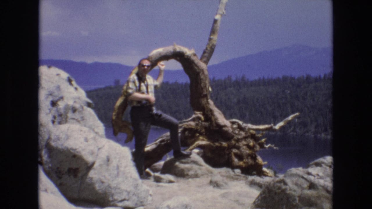 1973 Lake Tahoe home-movie frame showing a man in a plaid shirt and sunglasses posing on pale rocks beside a twisted root formation with forested mountains and water behind him.