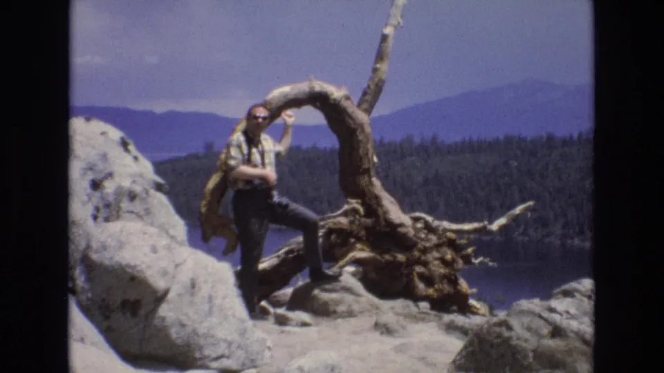 1973 Lake Tahoe home-movie frame showing a man in a plaid shirt and sunglasses posing on pale rocks beside a twisted root formation with forested mountains and water behind him.