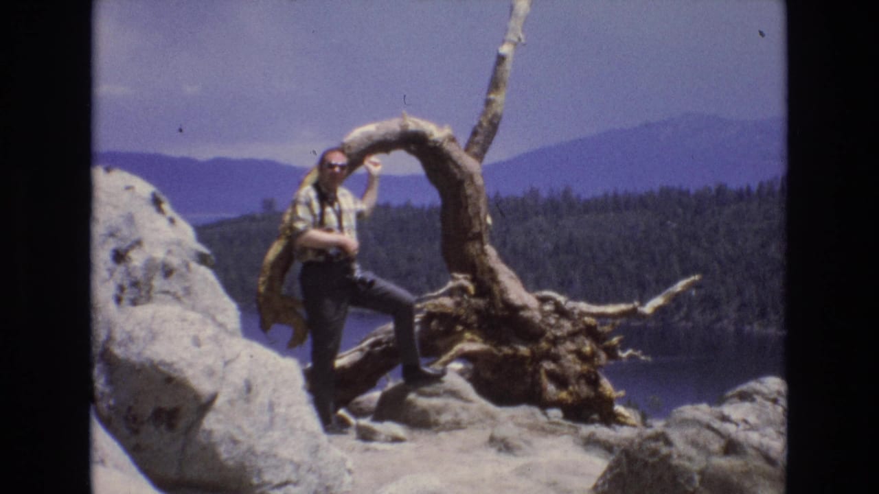 Man posing by the same twisted root and rocky lakeside overlook with mountains behind him.