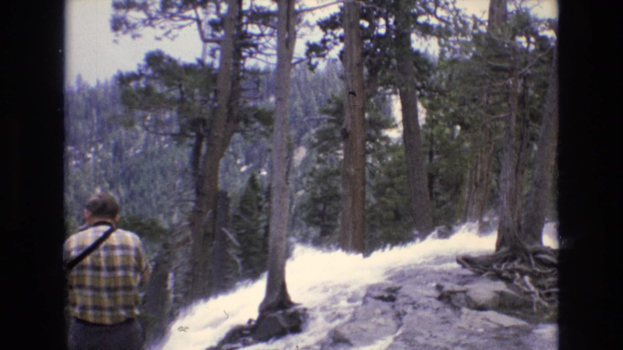 In documented 1973 Lake Tahoe region 8mm footage, a man seen from behind stands on a rocky ridge among pines while looking toward a cascading waterfall below.