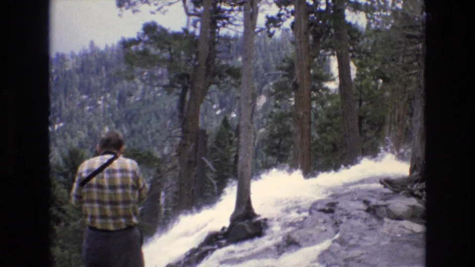 The man remains still on the rocky overlook in the Lake Tahoe region, framed by tall pine trunks and pale rushing water descending through the forested terrain.
