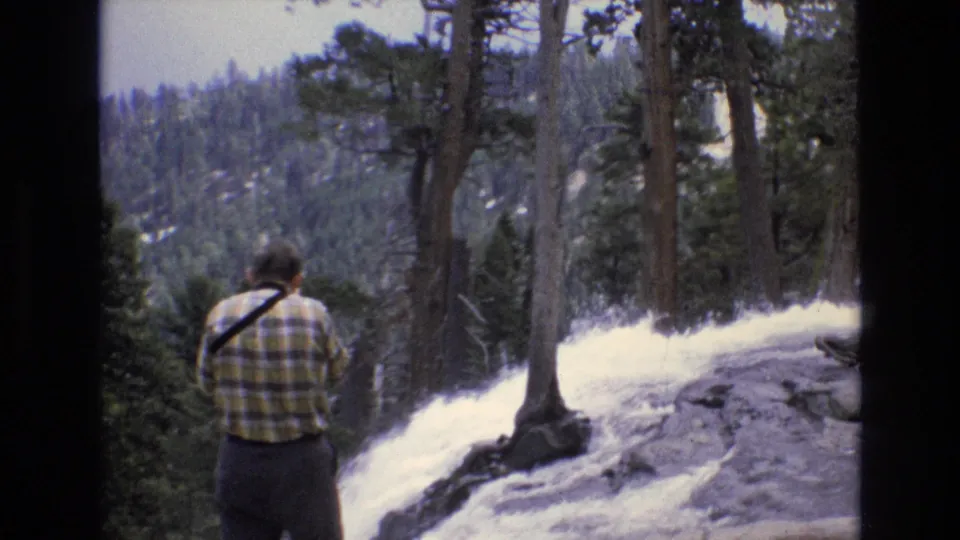 A rear view shows the visitor in a plaid shirt watching or filming the waterfall as pines and distant wooded slopes fill the 8mm home-movie frame.