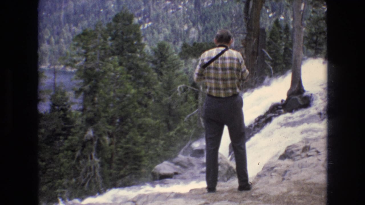 Adult man in a yellow-green plaid shirt stands with his back to the camera on a rocky Lake Tahoe area overlook near a waterfall and dense pine trees, filmed in 1973 8mm home-movie style.