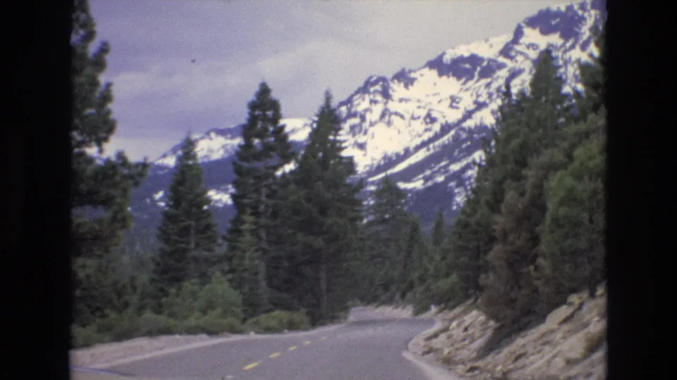 A winding two-lane road in the Lake Tahoe area curves through evergreen forest with snow-capped mountains visible under a cloudy sky.