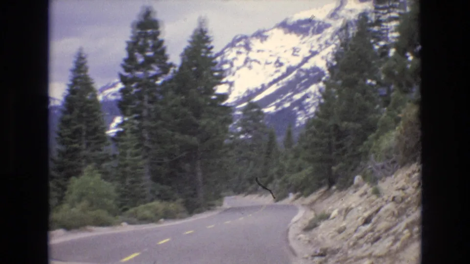 The road bends through conifer forest and rocky roadside terrain, with snow-covered Lake Tahoe area mountains rising in the background.