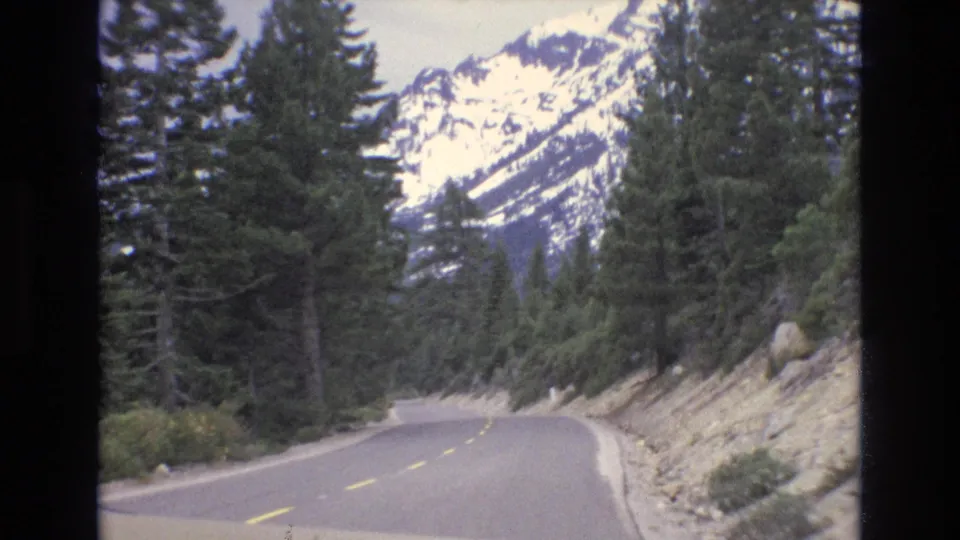 An empty paved road winds along a rocky shoulder through dense evergreens, with snow-capped mountains visible in the Lake Tahoe area distance.
