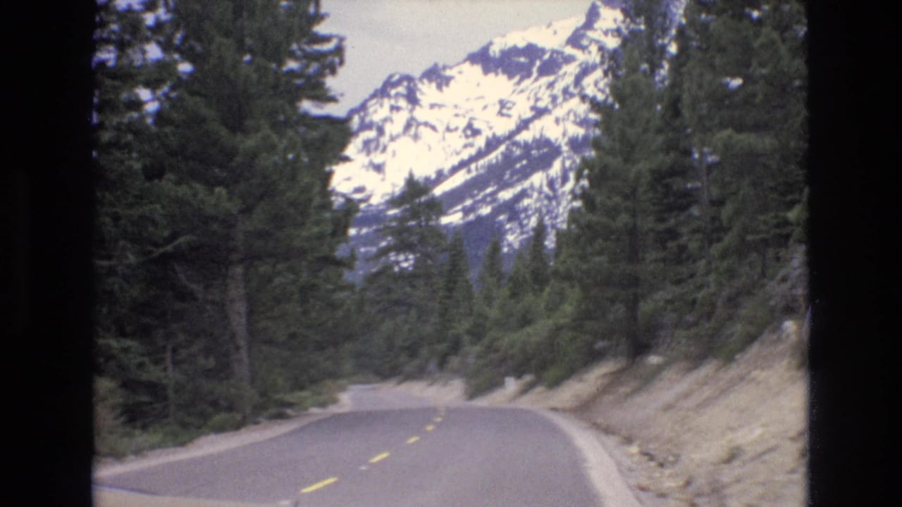 1973 Lake Tahoe, Nevada home-movie frame showing a two-lane asphalt road curving through evergreens toward snow-capped mountains.