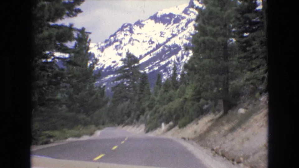 1973 Lake Tahoe, Nevada frame showing a forested highway curve with yellow lane markings and snow-covered mountains beyond the trees.