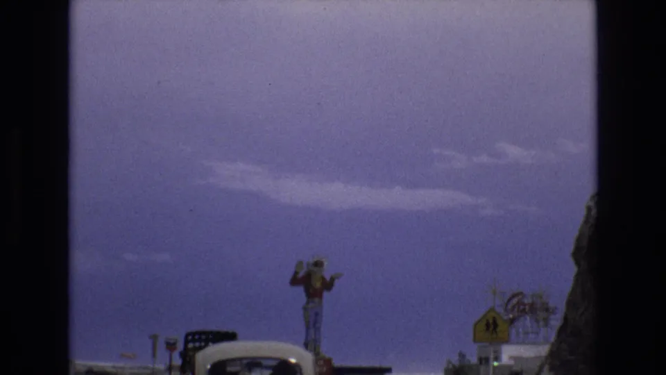 1973 Lake Tahoe, Nevada town approach showing a large cowboy figure above roadside signs, a car roof, and commercial structures against a purple-tinged sky.
