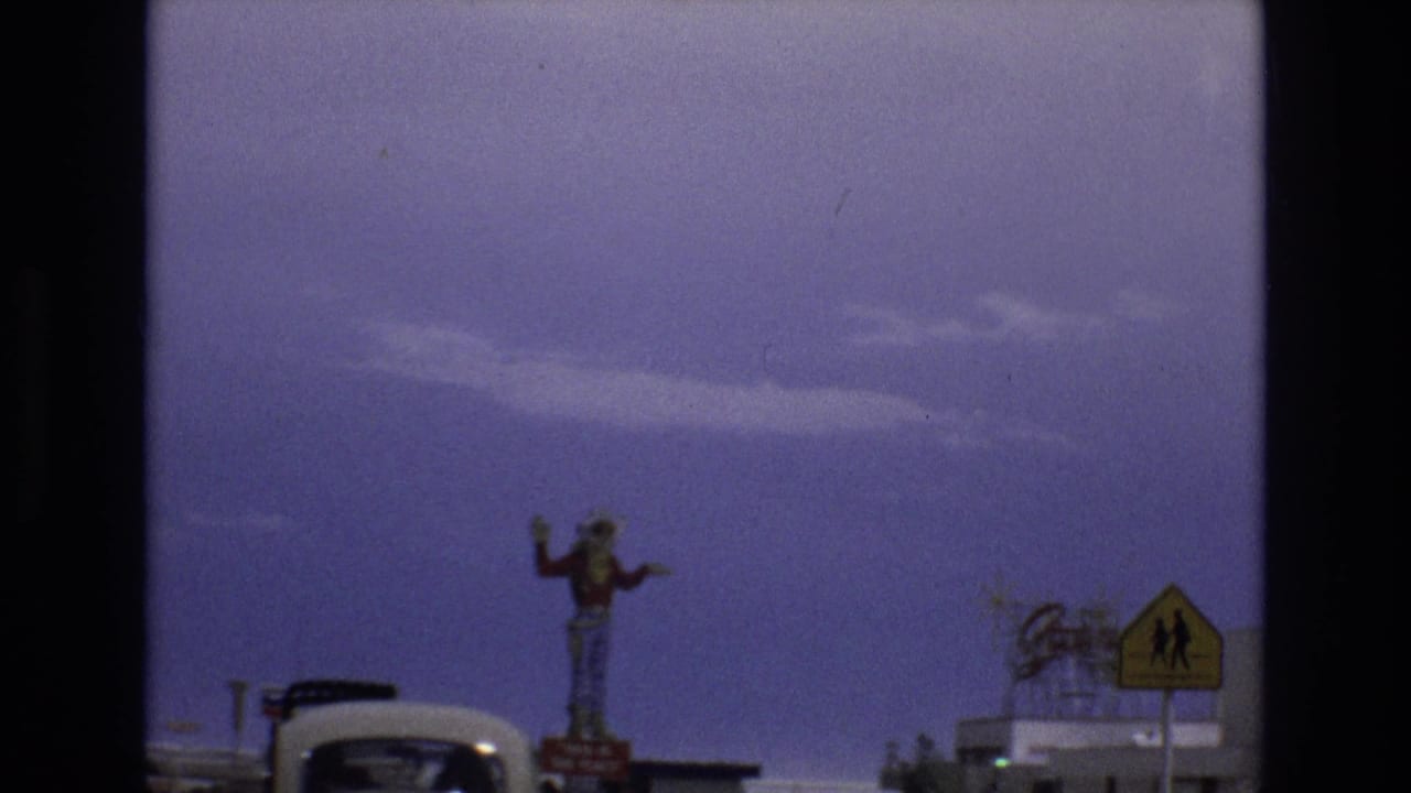 1973 Lake Tahoe, Nevada home-movie frame showing a tall waving cowboy statue near roadside buildings, with a Volkswagen Beetle and a yellow road sign partly visible.