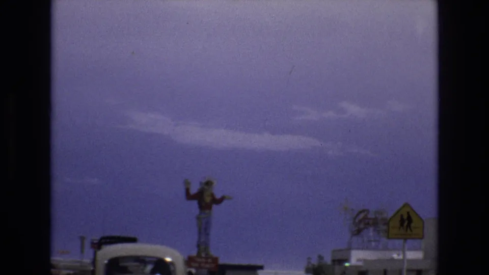1973 Lake Tahoe, Nevada home-movie frame showing a tall waving cowboy statue near roadside buildings, with a Volkswagen Beetle and a yellow road sign partly visible.