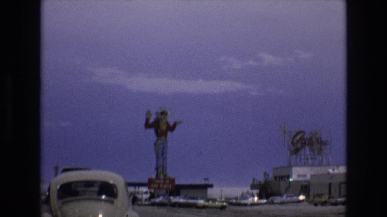 Tall cowboy statue above a roadside building with cars and a roof sign nearby.