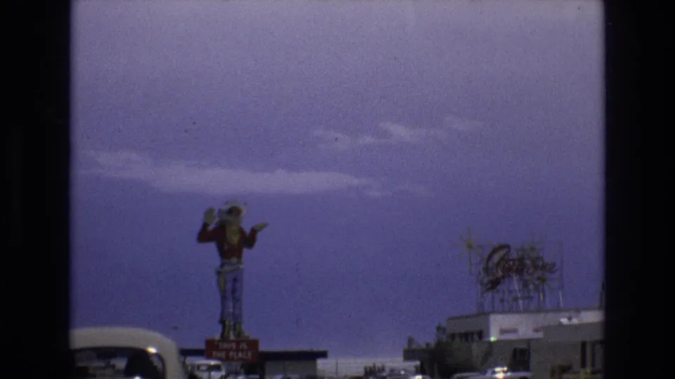 1973 Lake Tahoe, Nevada home-movie still showing the cowboy statue slightly left of center, a Beetle at the lower left edge, and a roof sign over a low building.
