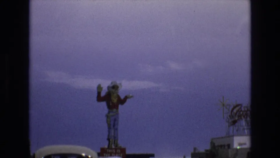 1973 Lake Tahoe, Nevada frame placing the waving cowboy statue against a purple-tinted sky, with a roadside building and roof sign at the right edge.