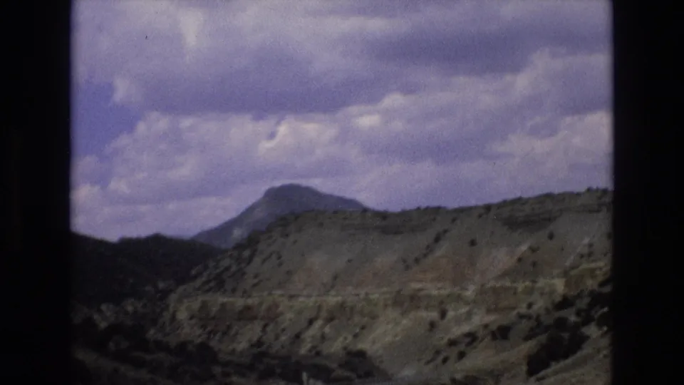 1973 8mm travel frame from the Lake Tahoe, Nevada area showing a paved road below dry layered hills, sparse scrub, a distant peak, and heavy clouds.