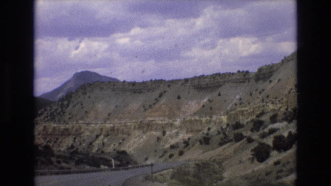 Roadside view of dry hills, sparse shrubs, a distant mountain, and a cloudy sky.