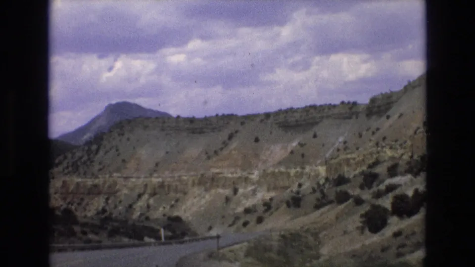1973 Lake Tahoe, Nevada travel footage frame showing a roadside passage through dry hills with scattered shrubs, a distant mountain form, and a purple-tinted cloudy sky.