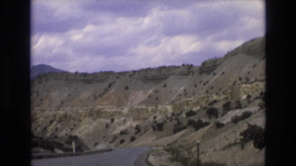 1973 8mm frame from the Lake Tahoe, Nevada area showing a curving paved road at the base of scrub-covered slopes beneath a cloudy sky.