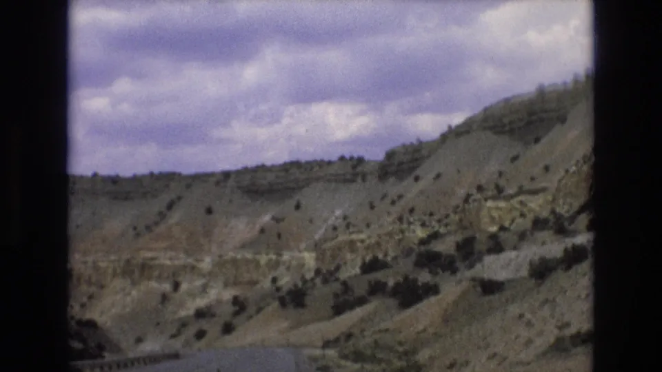 1973 Lake Tahoe, Nevada area frame showing the edge of a paved road with guardrail below dry layered hills, scattered shrubs, and a broad cloudy sky.